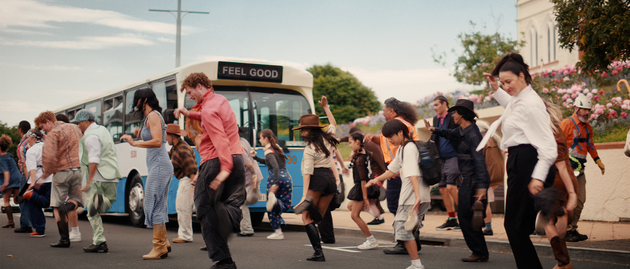 A large group of happy people dancing in the street. A bus with the words "Feel Good" in the background.